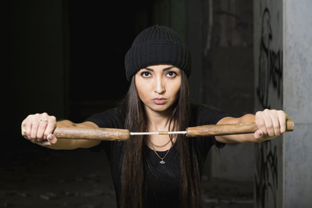 Fighter girl, a brunette in a black hat, stands in an old building. In front of him he is holding a Chinese weapon - nunchucks. Looks into the cameraの写真素材