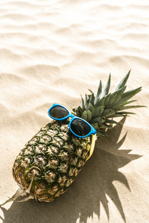 A funny photo of a pineapple with sunglasses lying on a sandy beach