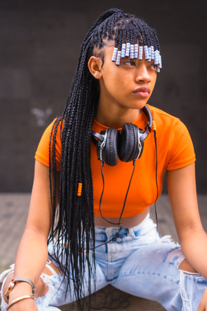 Lifestyle with a young trap dancer with braids in the city. Black Race Girl African Ethnicity With Orange T-Shirt And Cowboy Pants, Vertical Photoの写真素材