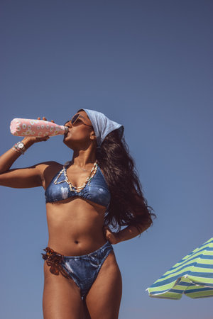 A black girl standing on the beach while she drinks water with sunglasses on on a sunny day with a blue background. Summer vibes concept 2021.の写真素材