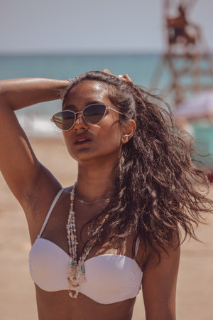 A happy, smiling black girl on the beach as she pulls her hair into a ponytail on a sunny day. Summer vibes concept 2021.の写真素材
