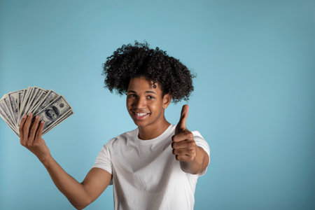 Young latin man with afro hair holding new one hundred dollar bills smiling happy and positive,...