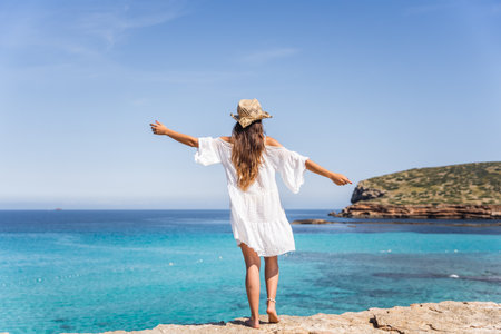 Girl with white dress and brown hat walking on a paradise beach with crystal turquoise water in the backgroundの写真素材