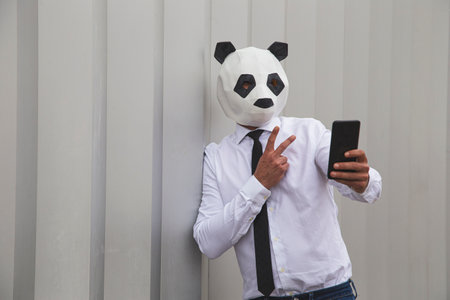 Man in white shirt, tie and panda mask taking smartphone selfie against wallの写真素材