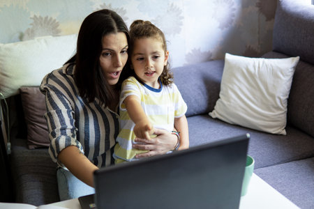 Young white Caucasian woman working from home with her son in her arms, with astonished face, pointing at the laptop screenの写真素材