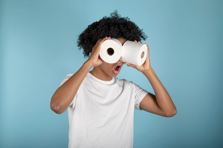 Funny young man with afro hair looks through toilet paper rolls, simulating binoculars. Isolated on blue background. Studio portrait.の写真素材