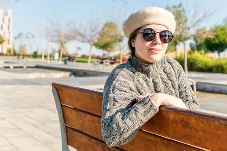 Young Caucasian woman, with round sunglasses and beret, looking at camera, sitting in a park, on a sunny day.の写真素材