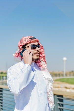 Arab man talking on the phone, in traditional dress, at park. Vertical shot.