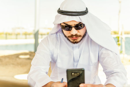 Arabian man, in traditional cloth, seating at park, checking his mobile phone.の写真素材