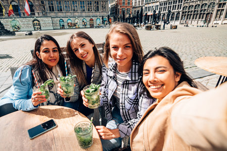 selfie time. Happy four smiling girlfriends sitting outdoor with cocktails taking a group photo. selective focus on Indian left woman face.の写真素材