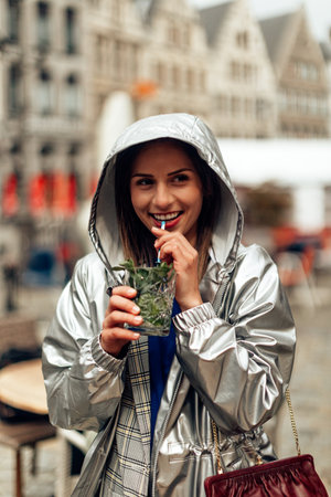 cheerful smiling young woman in k-way drinking a cocktail on a rainy day in a European city. tourist in Antwerpen, Belgiumの写真素材