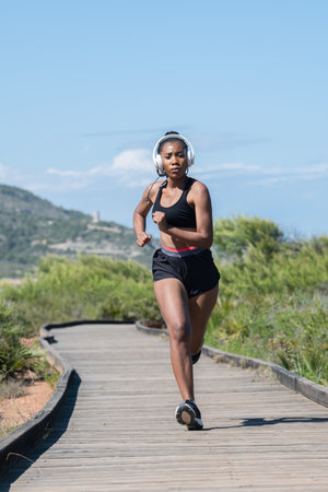 Vigorous afro american woman using headphones and sports clothes running towards the camera: Selective focus. Exercise and healthy lifestyle concept.の写真素材