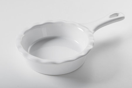 From above of new empty clean round white ceramic baking bowl with handle and wavy edges on white...