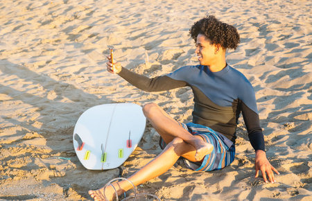 African American man sitting on the sand with surfboard takes a photo with a smartphoneの写真素材