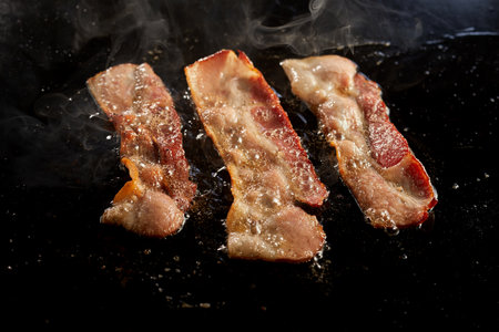High angle of delectable bacon strips cooking in oil on hot black surface