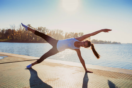 young woman in leggings and white undershirt   on pontoon at lake practice yoga, sunny autumn day, water droplets all around from near by water fountainの写真素材