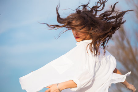 young woman in white shirt with hair in motion, outdoor spring day, blue sky in backgroundの写真素材
