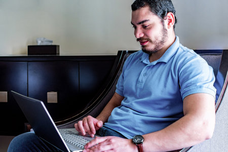 Young freelancer man using laptop, sitting in an armchair, working remotely.