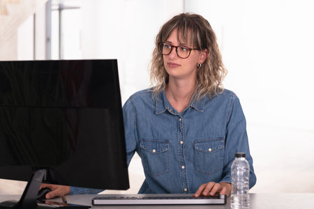 Female worker wearing casual jeans shirt and eyeglasses in the office using computer on an illuminated background.の写真素材