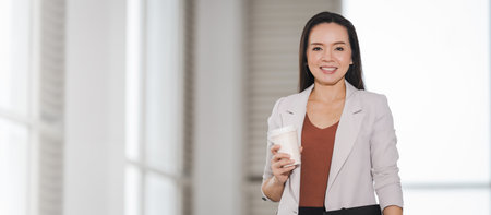 A happy successful Asian businesswoman holding a takeaway coffee cup, document file and digital tablet in the business buildingの写真素材
