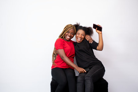 two happy and excited young black women sitting together, holding their phones, advertising conceptの写真素材