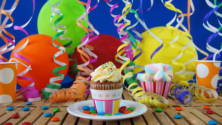 Cupcake on rustic wooden table with background of colorful balloons, plastic cups and candies with blue wall in backgroundの写真素材