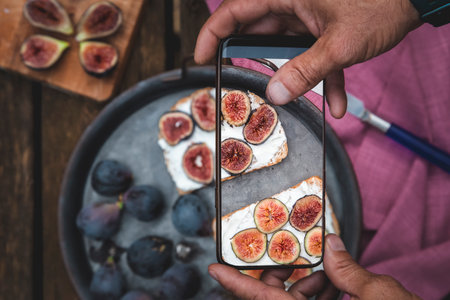 Man hands takes picture of food on the table with the phone. Breakfast. Sandwich with soft ricotta and figs. Smartphone photo for social media or blog post. Vegetarian, healthy, organicの写真素材