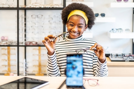 Young woman african american entrepreneur is fashion vlogger show glasses to cellphone and smiling selling it online live streaming in eyeglasses shopの写真素材