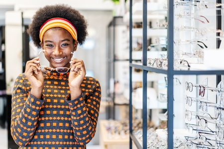 Need a new glasses. Young woman african american in optic store choosing a new eyeglasses frame. Medical, health care concept, used correct or assist defective eyesightの写真素材