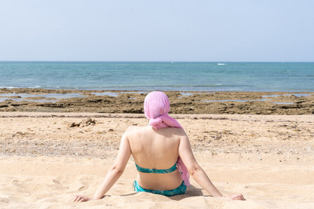 Woman sitting on the beach looking at the sea with pink scarf on her head, cancer.の写真素材