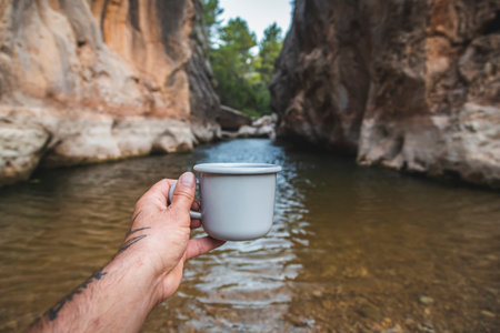 Hands of male tourist standing on the river bank and holding a mug with hot drink.の写真素材