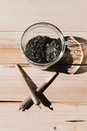 Overhead shot of a jar with crushed cannabis and two crossed joints on a wooden table. Concept of therapeutic cannabis, medical cannabis, legal cannabisの写真素材