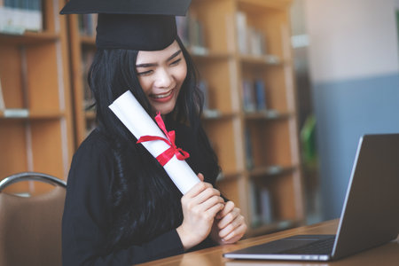 A young Asian female university graduate expressing joy and excitement to celebrate her achievement of degree graduation in front of a laptop making a remote video call to her parents at homeの写真素材