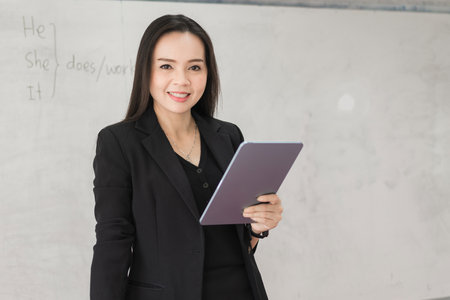 Adult confident cheerful Asian woman teacher in black business suit uniform with digital tablet and laptop teaching modern in classroomの写真素材