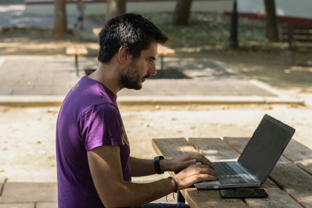 young man working at his computer on a wooden table in the park on a sunny dayの写真素材
