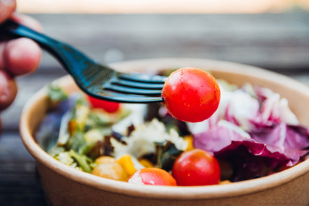 Close-up of a hand with a fork and a tomato. In the background a mediterranean salad in a cardboard container.の写真素材