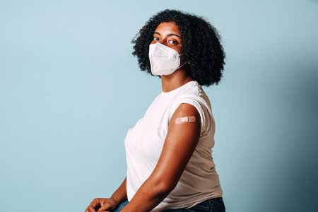 young afro american woman wearing face mask showing her vaccinated arm on blue background.の写真素材