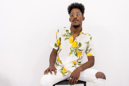 A young Cuban man with curly hair sitting in a white flowered shirt and glasses. Posed looking at...