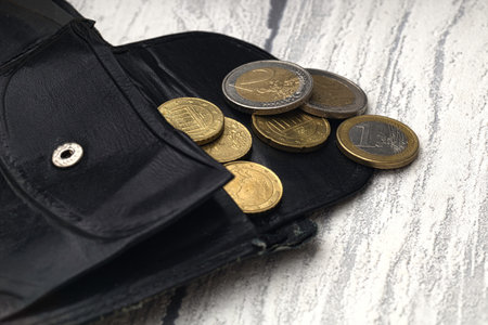 some euro coins pour out of a old use black leather wallet on a white wooden background (selective focus on coins)の写真素材