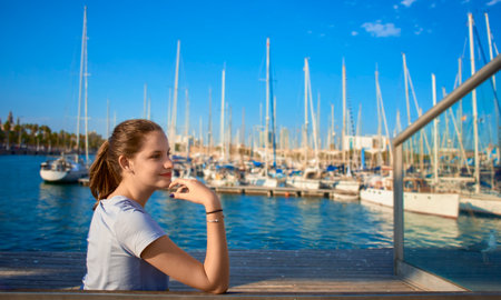 A girl sitting on a pier on the foreground in the afternoon with a background with boats out of focus. Blue sky and water tooの写真素材