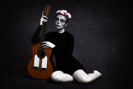 Woman with her face painted like a Catrina and flowers in her hair on dark background holding a guitar. Day of the Deadの写真素材