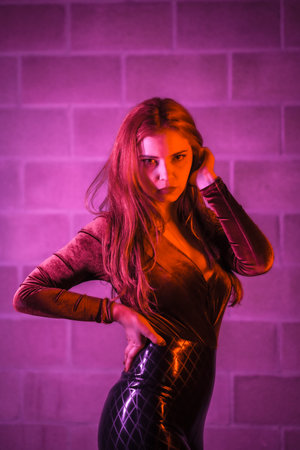 Red-haired girl in latex, brown and black tight suit on a brick wall, posed at night illuminated...