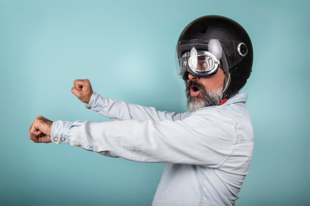 Profile of bearded hipster in denim shirt with helmet and glasses pretending to ride a motorcycle, isolated on blue studio wall.の写真素材