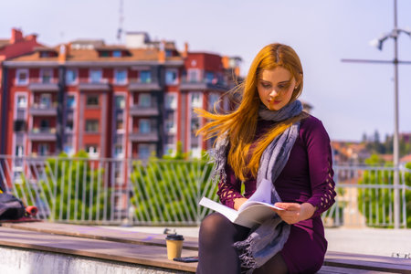 Beautiful woman in purple dress and scarf reading a book in a park in the city, imagining and enjoying readingの写真素材