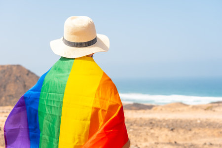 Posing of an unrecognizable gay person with a gray t-shirt and with the LGBT flag by the sea, symbol of homosexualityの写真素材