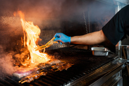 Hand with glove of a professional cook covering with spices on a piece of meat on the grill with a big flame coming out of the grill.の写真素材