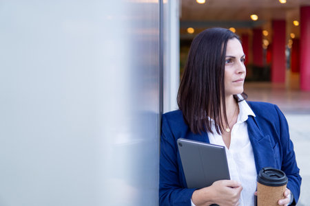 Beautiful Caucasian businesswoman with a cup of coffe and tablet in the street outside of an office buildingの写真素材