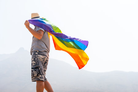 An unrecognizable gay person in a gray t-shirt and white hat waving the LGBT flag in a cloudy sky, symbol of homosexualityの写真素材