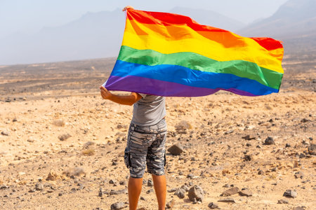 A gay person in a gray t-shirt and black cap with the LGBT flag waving in a desert with the wind, a symbol of homosexualityの写真素材
