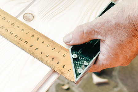 Close-up of craftsman hands in measuring wooden plank with angular ruler and pencil. Woodwork...
